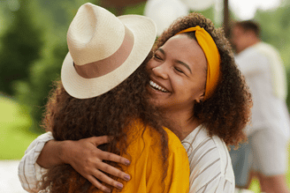 A person wearing a yellow headband and a striped shirt hugs another person wearing a straw hat and a yellow shirt.