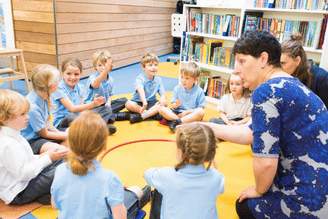 A teacher leads a group of young students in a circle on a yellow mat in a classroom with bookshelves in the background.