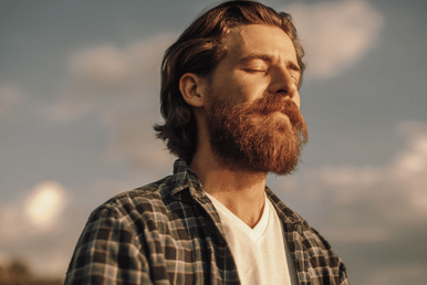 A person with a beard and closed eyes wears a plaid shirt and a white t-shirt.