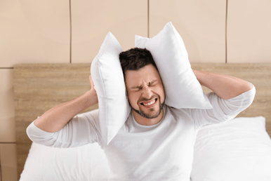 A man sits on a bed with white pillows covering his ears, his eyes squeezed shut in discomfort.