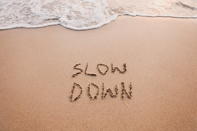 The words "SLOW DOWN" are written in the sand on a beach, with a gentle wave washing ashore in the background.