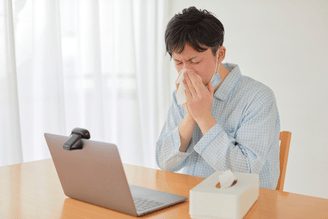 A person wearing pajamas blows their nose into a tissue while sitting at a table with a laptop and a box of tissues.