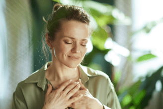 A woman with her eyes closed places her hands over her chest, with lush green plants in the background.
