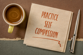 A yellow mug of coffee sits next to a folded napkin with "PRACTICE SELF COMPASSION" written in red ink, and a silver pen lies to the right.