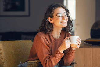 A smiling person wearing glasses and a rust-colored sweater holds a mug of coffee while sitting in an armchair.