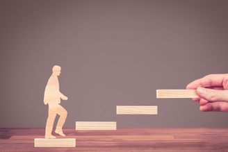 A wooden silhouette of a person climbing a staircase made of wooden blocks, with a hand adding another block to the top step.