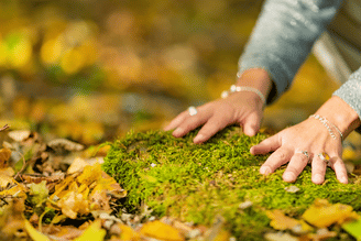 Hands with rings and bracelets rest on a mossy surface surrounded by fallen autumn leaves.
