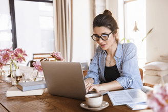 A person wearing glasses and a striped shirt works on a laptop at a wooden table with flowers and a coffee cup.