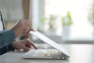 Person's hands closing a laptop on a white desk with a blurred background of plants and a window.