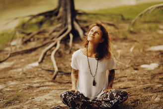 A person sits cross-legged on the ground in front of a tree with exposed roots, looking upwards.