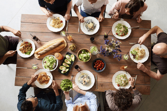 A group of people sitting around a wooden table, enjoying a meal with various dishes including pasta, salad, bread, and avocado.