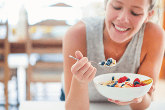 A person smiling while holding a spoon with blueberries and cereal, and a bowl of cereal with strawberries and blueberries.