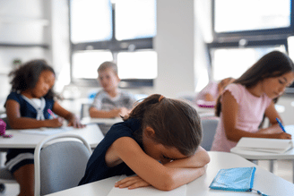 A young student rests her head on her arms on a desk in a classroom while other students work.