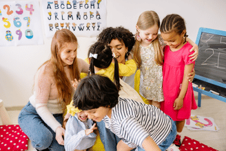 A teacher and several children are gathered together in a classroom setting, interacting joyfully. Alphabet and number charts are visible on the wall behind them, and a chalkboard is to the right.