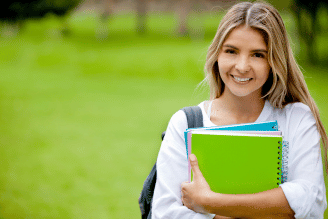Student standing outdoors on a green campus lawn, smiling while holding a stack of colorful notebooks and a backpack strap over one shoulder.
