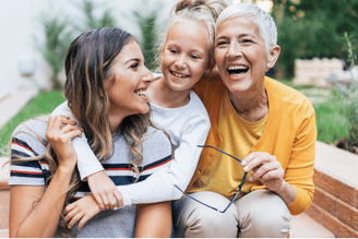 Three people smiling outdoors: an adult with a child on their lap and an older woman in a yellow shirt, sitting near a brick wall with greenery in the background.