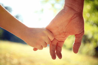 Two hands reaching and holding each other outdoors, with sunlit greenery in the blurred background.