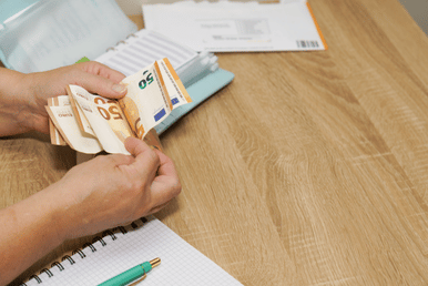 Hands counting and holding several banknotes at a wooden table, with a notebook, pen, and paperwork in the background.