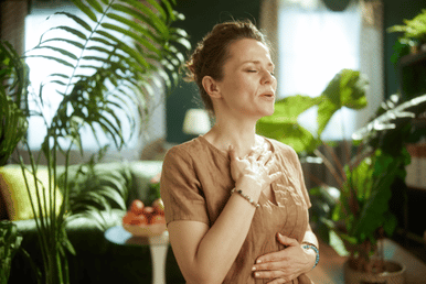 A person in a brown dress stands indoors among many green potted plants, holding a small bouquet and smiling while looking to the side.