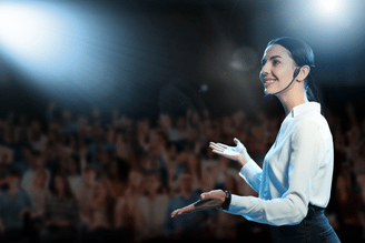 Performer singing and gesturing on a brightly lit stage with a cheering crowd in the background at a concert.