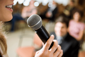 Close-up of a hand holding a silver microphone near a speaker’s mouth, with a softly blurred audience seated in a conference or event room.