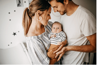 A couple in a cozy room holding a baby and looking at each other, with the baby in a striped swaddle between them.