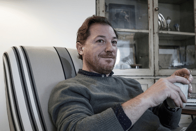A man with short hair and a beard lounges in a striped armchair, leaning back with arms resting on the chair beside a window and shelf.