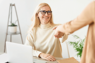 A woman with glasses sits at a table using a laptop while a person in the foreground extends a hand to hand over a parcel, with a plant and bright office background behind her.