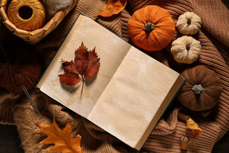 An open cream-colored book on a burlap cloth surrounded by autumn leaves, small pumpkins, and walnuts, with decorative gourds and acorns nearby on a dark wooden surface.