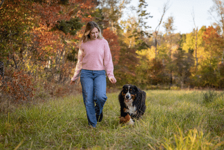 Person in a pink sweater walking on a grassy path with a black-and-brown dog in an autumn park with trees showing orange and yellow leaves.