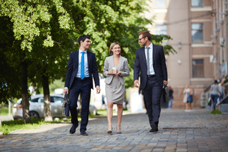 Three business professionals walk along a tree-lined sidewalk beside brick buildings, with one woman in a light dress and two men in suits moving toward the camera.