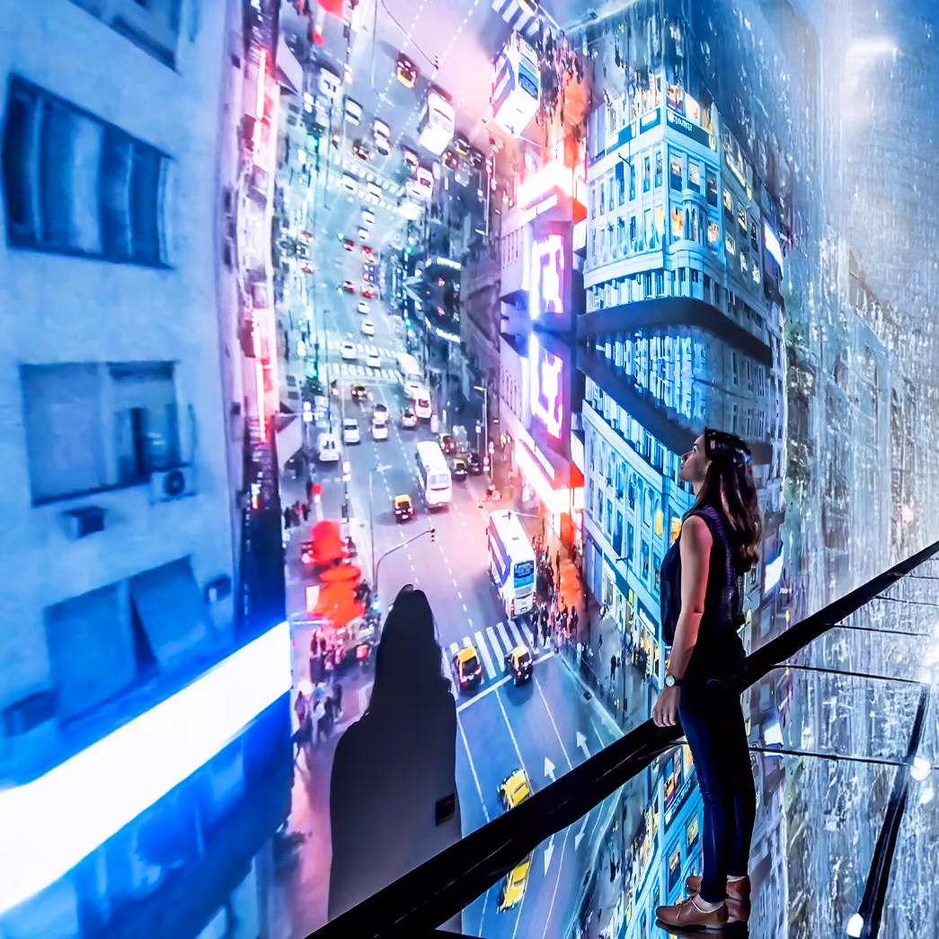 Visitor on a walkway inside an immersive room, surrounded by floor-to-ceiling video of a city street seen from above