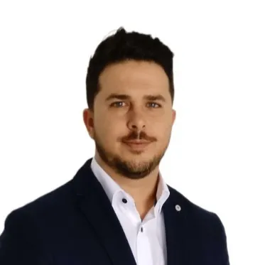 Portrait of a man with short dark hair and a trimmed beard wearing a white shirt and dark blazer against a white background.