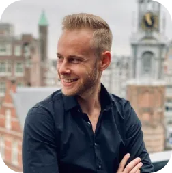 Smiling man with short blond hair wearing a black shirt, standing outdoors with blurred historic buildings in the background.
