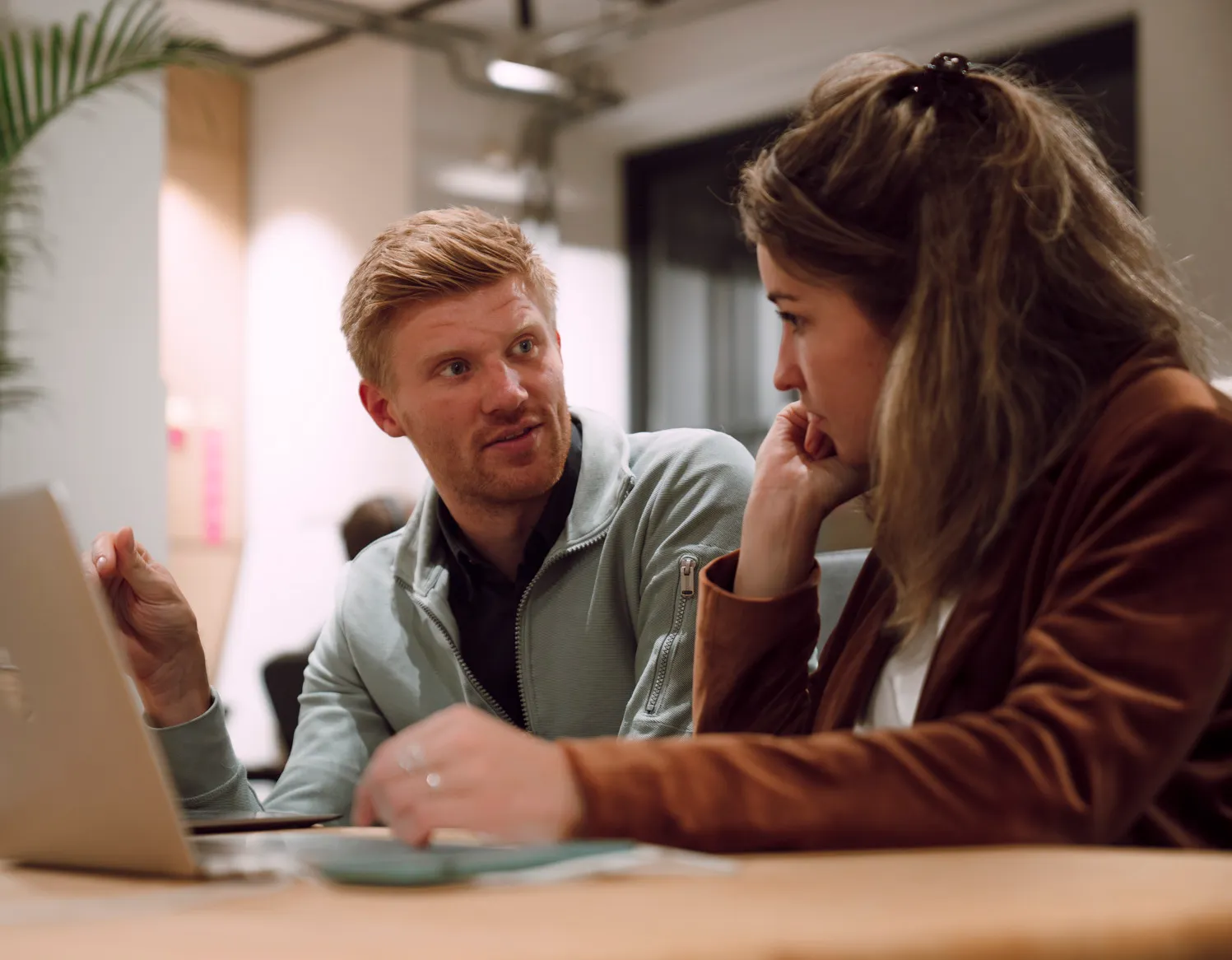 Two colleagues engaged in a focused discussion at a table with a laptop in an office setting.