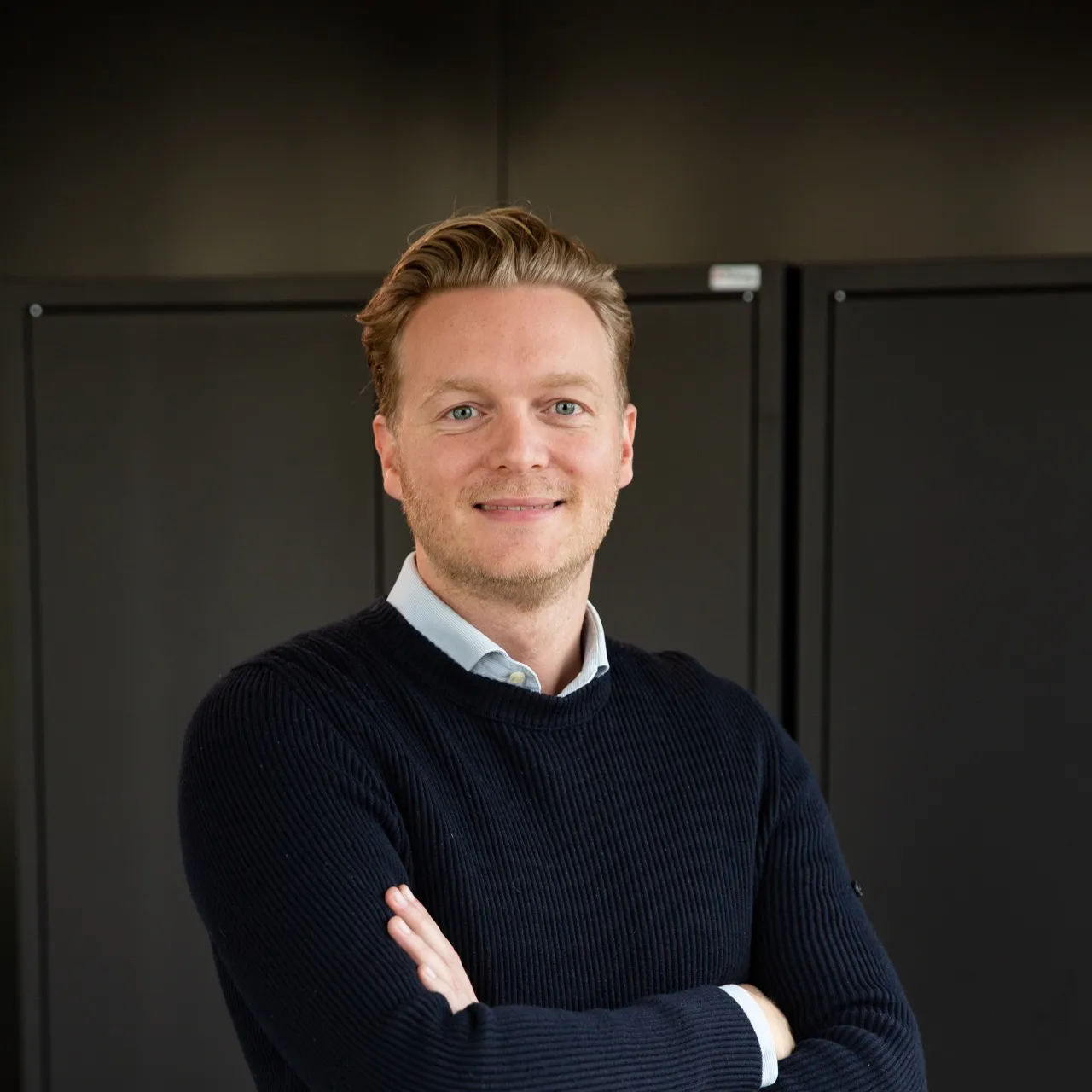 Smiling man with blond hair wearing a navy blue sweater and light blue collared shirt standing with arms crossed in front of dark cabinets.