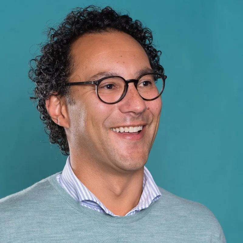 Smiling man with curly hair and black-rimmed glasses wearing a light blue sweater over a striped collared shirt against a teal background.