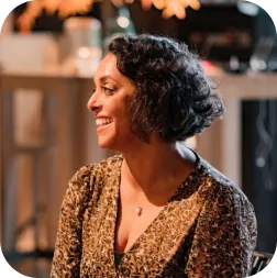 Woman with short curly hair smiling and looking to the side in a warmly lit indoor setting.