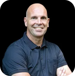 Smiling bald man in a dark blue polo shirt with folded arms against a black background.