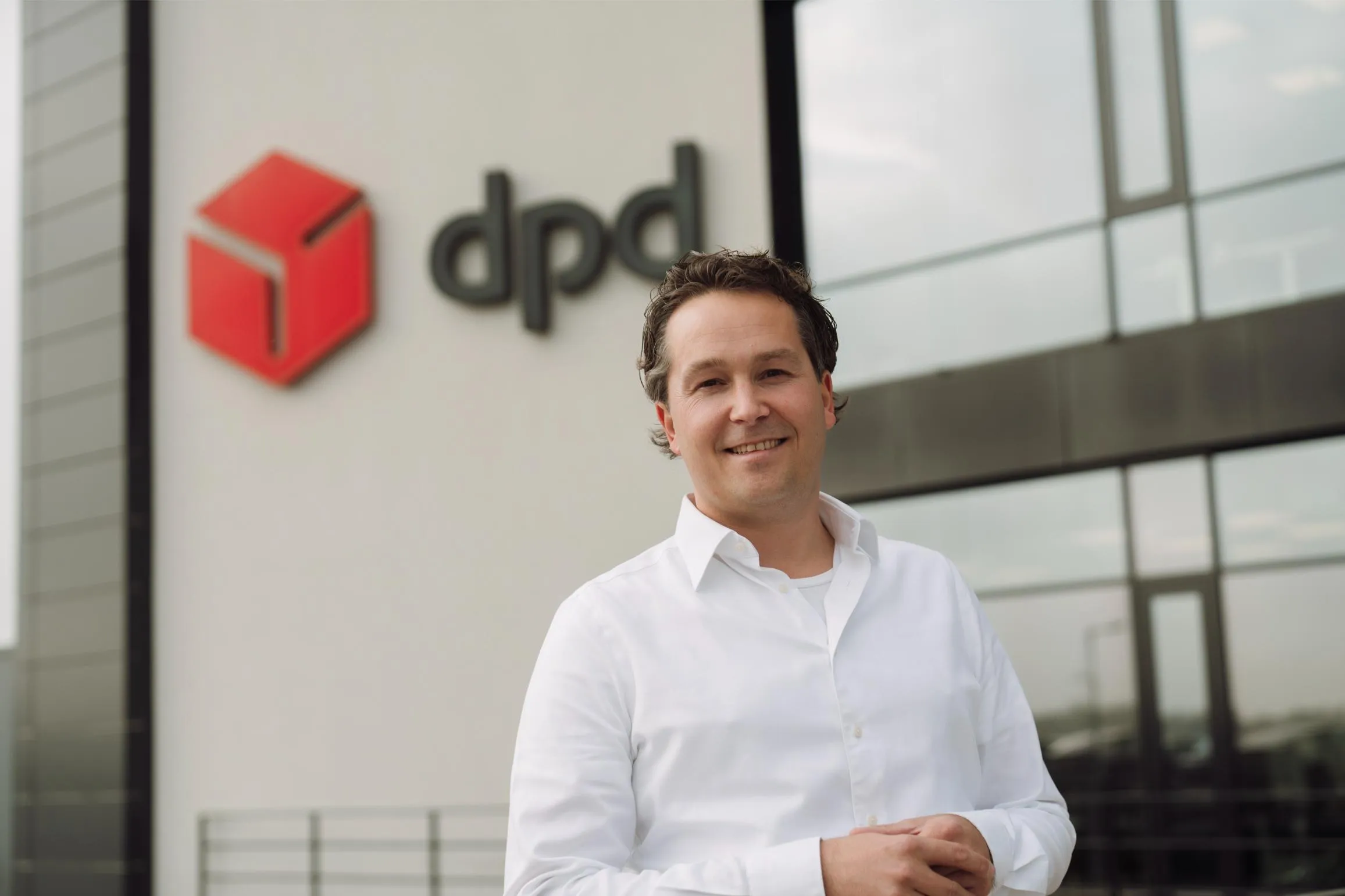 Smiling man in a white shirt standing outside a building with a red dpd logo on the wall.