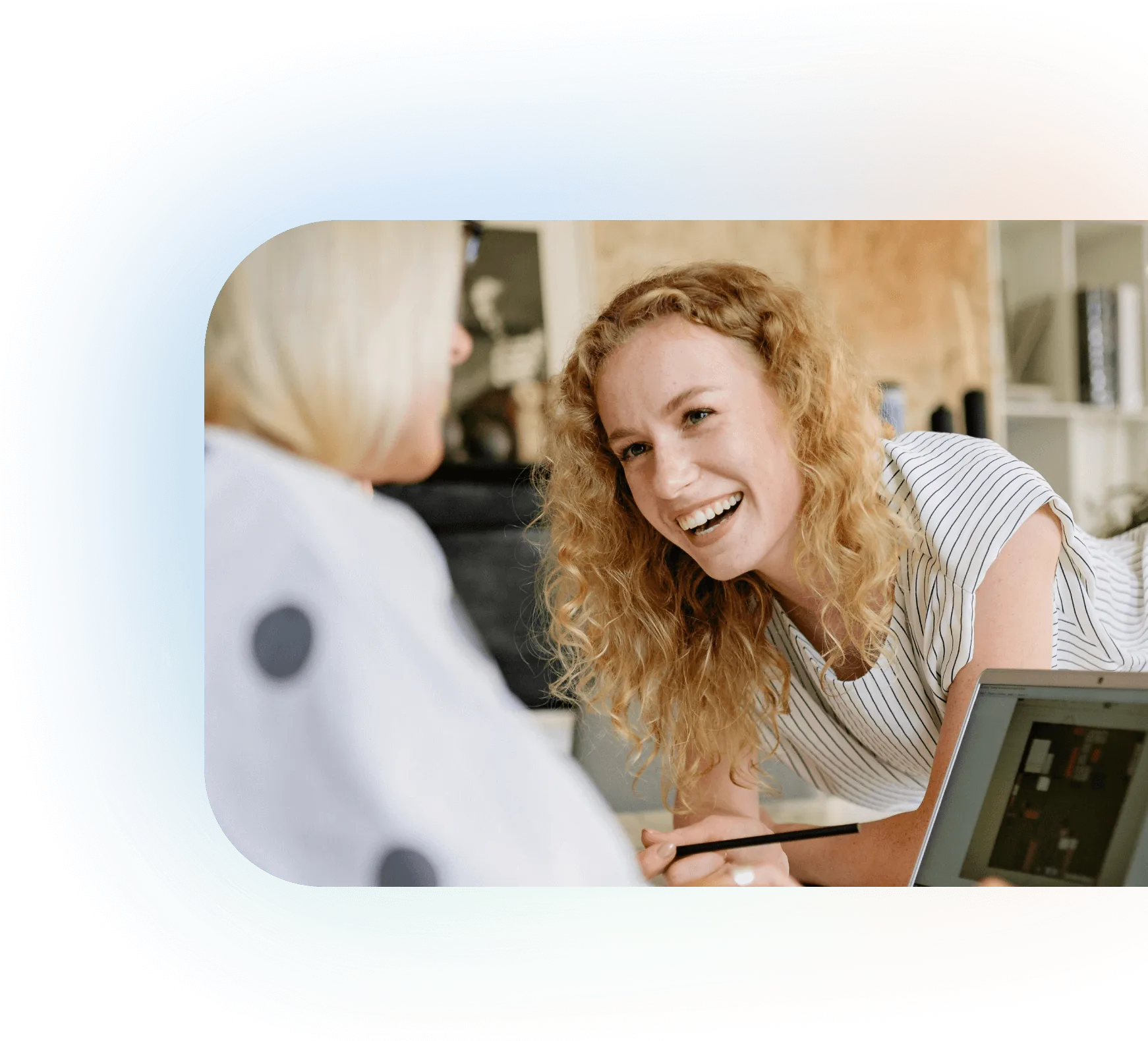 Young woman with curly hair smiling and holding a pencil while talking to another person in a casual office setting with a laptop nearby.