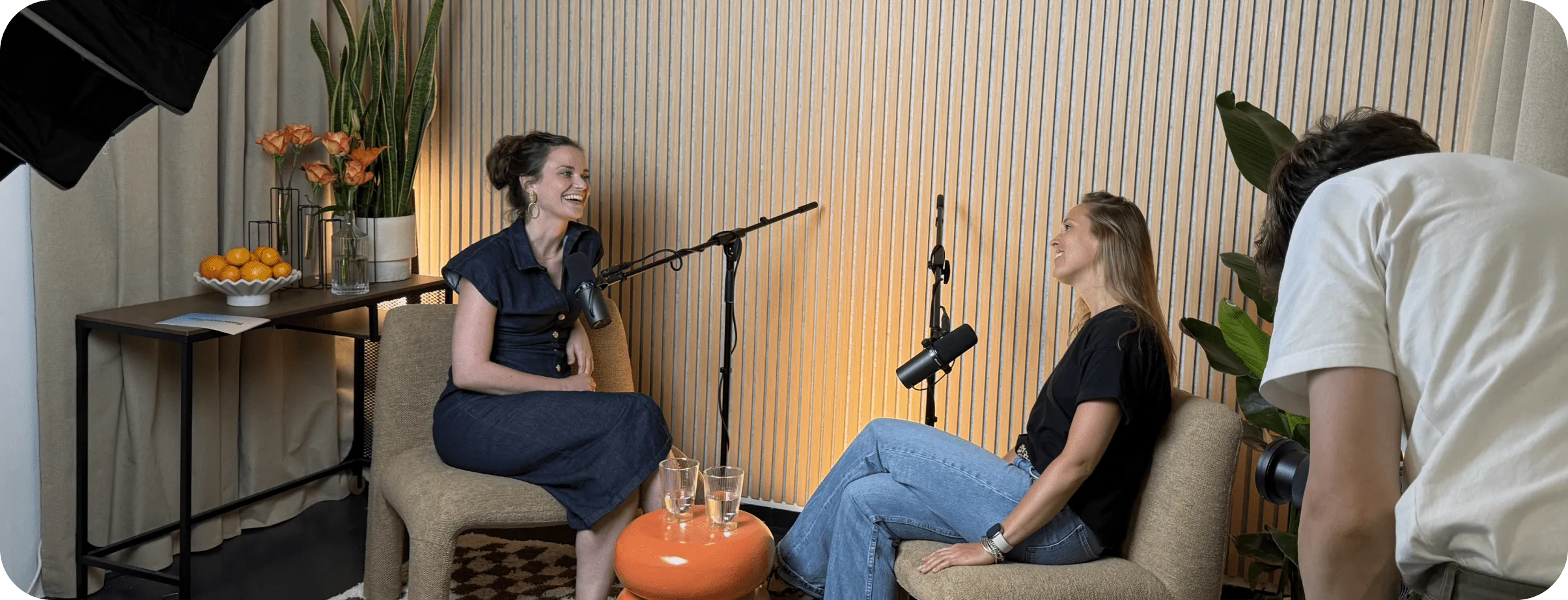Two women laughing and talking into microphones in a cozy podcast recording setup with a cameraman filming them.