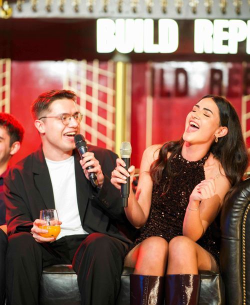 Two people sitting on a black couch. They are both dressed elegantly. The man is holding a microphone and a glass of drink. The woman is laughing.