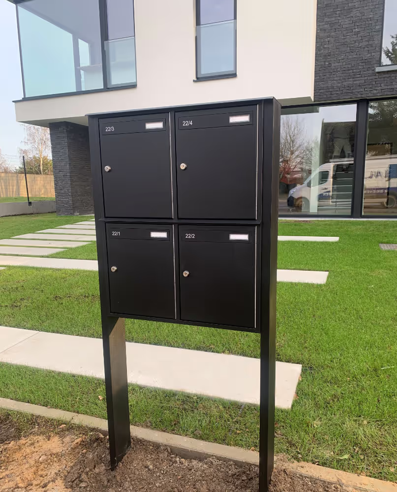 Een zwart brievenbusgeheel op staander voor een appartementsgebouw.