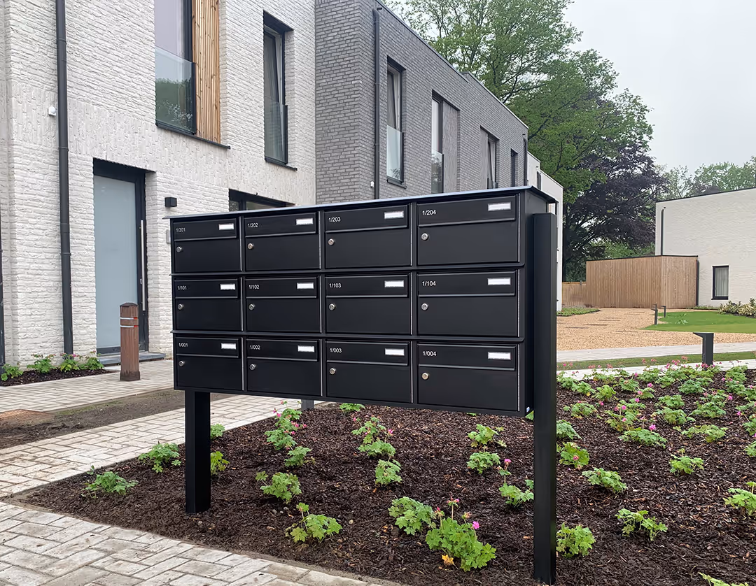 Een zwart brievenbusgeheel op staander voor een appartementsgebouw.