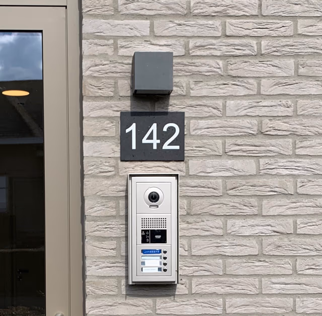 Un interphone contre un mur blanc, surmonté du numéro 142.