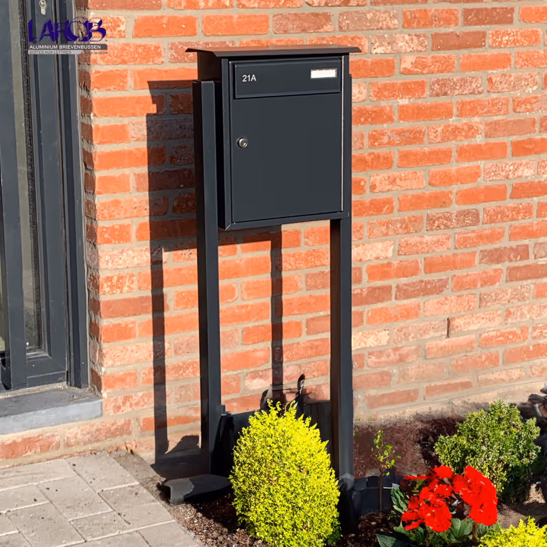 Een zwarte brievenbus op staander voor een bakstenen muur.