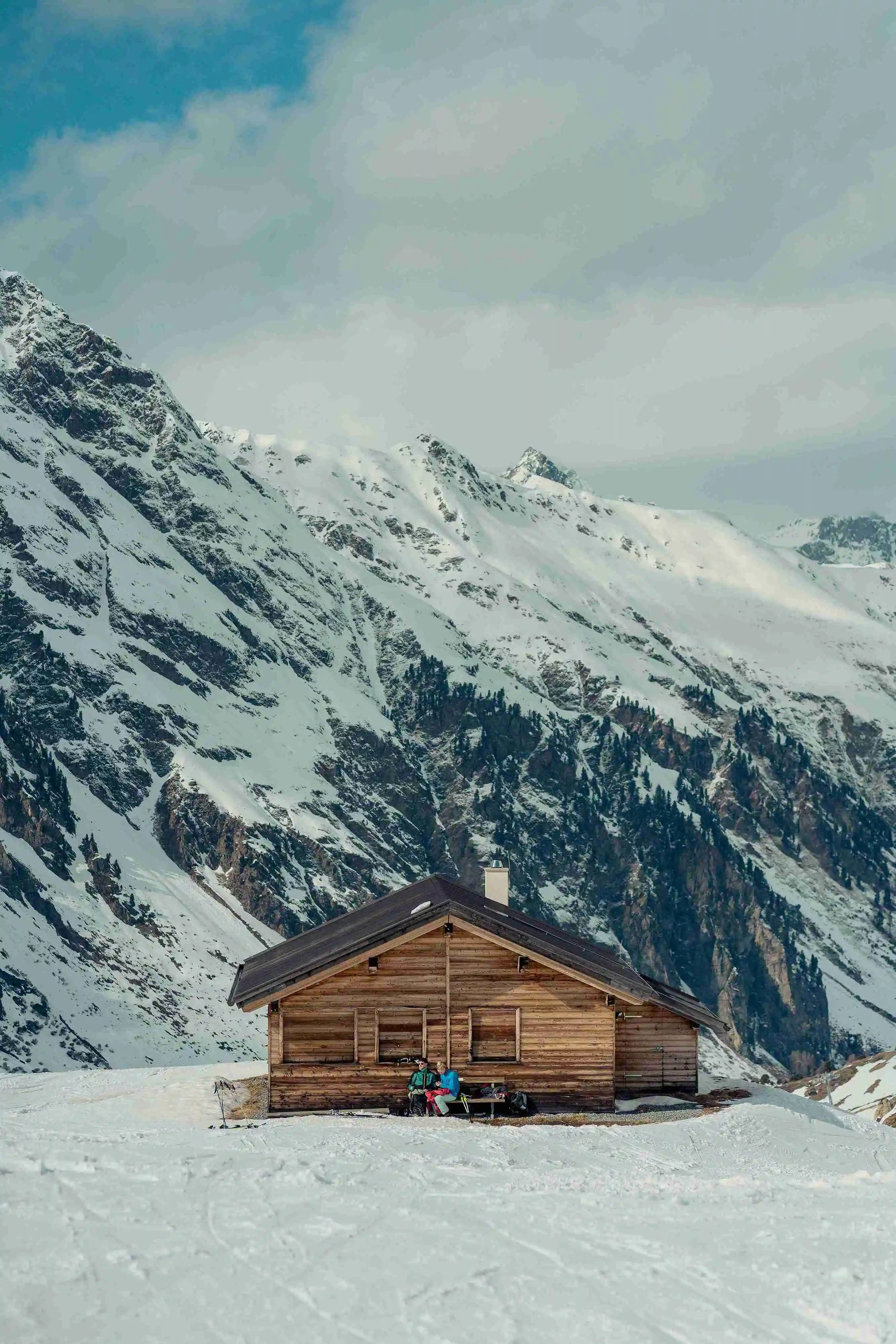 Eine holzverkleidete Berghütte steht inmitten einer schneebedeckten Landschaft, umgeben von majestätischen Bergen unter einem bewölkten Himmel. Ideal für Ferienwohnungen und Short-Term-Rentals, die eine ruhige Flucht ins Gebirge bieten.