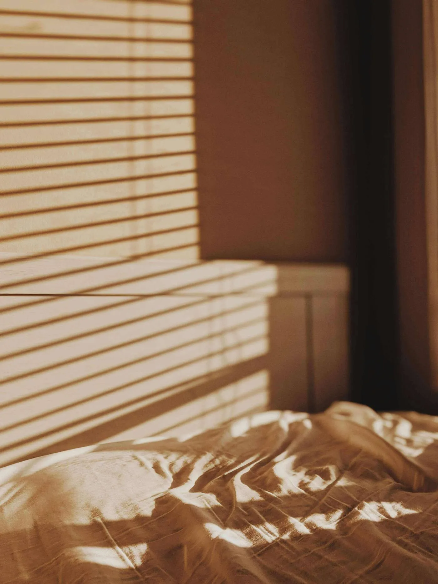 Das sanfte Sonnenlicht fällt durch die Jalousien und wirft gestreifte Schatten auf die unordentlich drapierte Bettdecke in einem gemütlichen Schlafzimmer. Das warme Licht schafft eine einladende Atmosphäre.