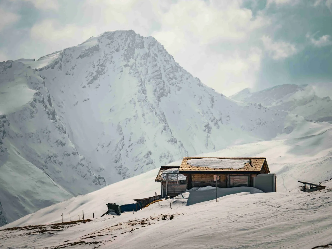 Eine gemütliche Berghütte aus Holz steht auf einer schneebedeckten Fläche. Im Hintergrund erheben sich majestätische Berge, die von Wolken umhüllt sind. Diese Szenerie eignet sich ideal für Ferienwohnung und Ferienvermietung im alpinen Stil.